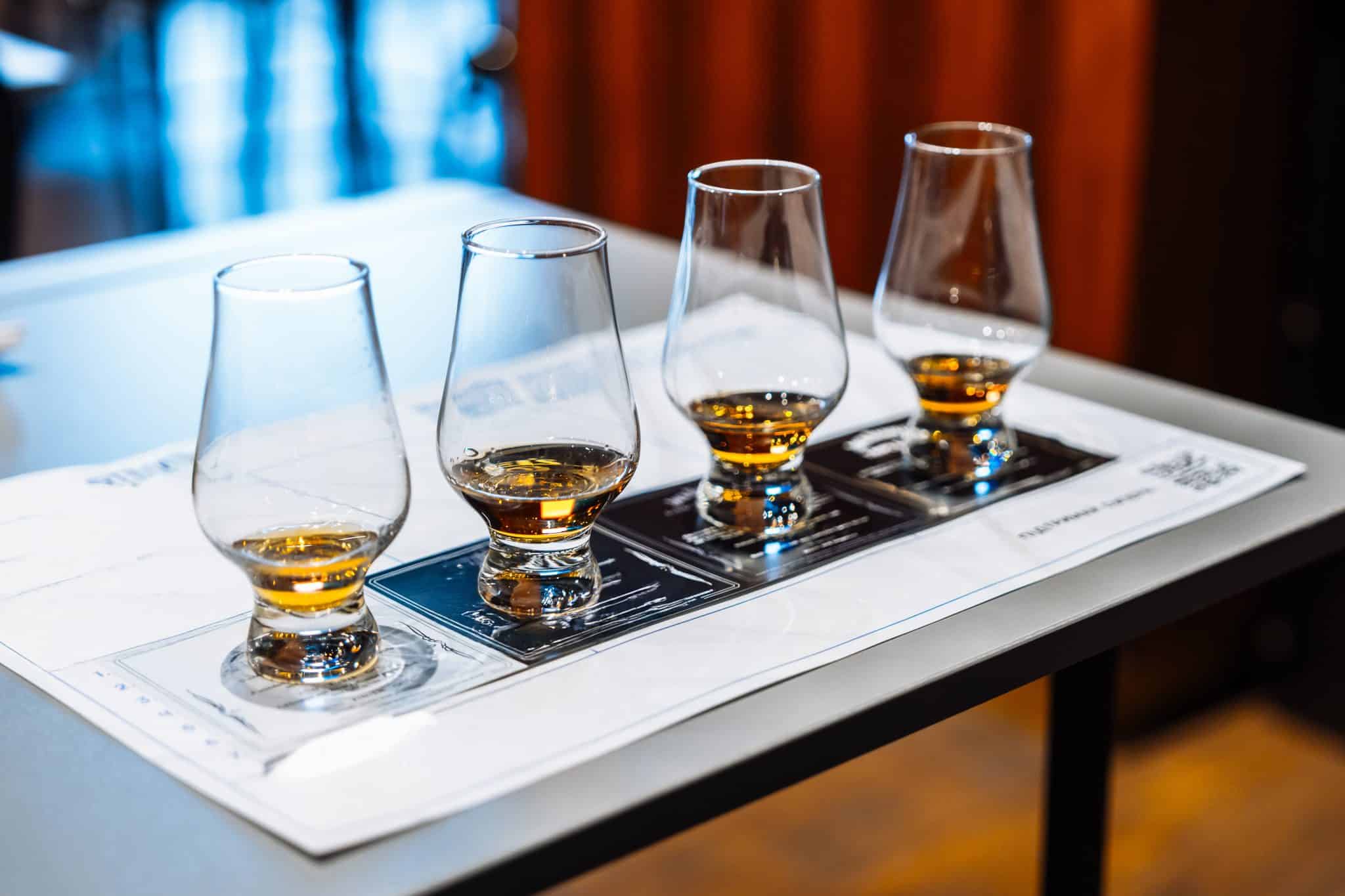 A group tastes whiskey from four glasses on a tasting mat indoors.
